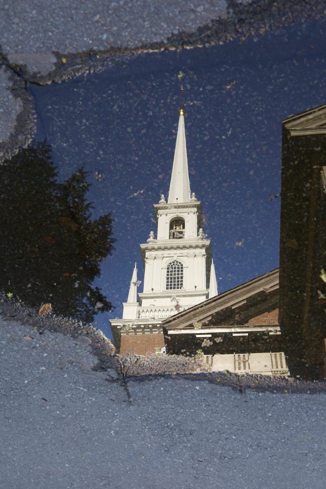 A church steeple and blue sky reflected in a puddle on the pavement.
