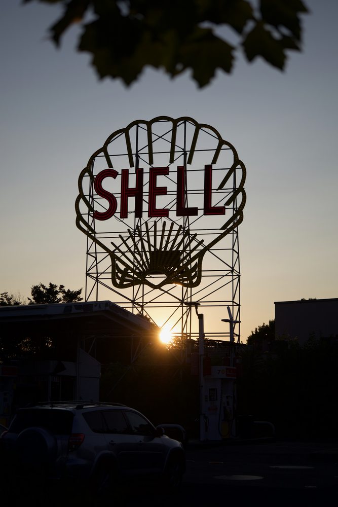 A large Shell sign silhouetted above parked cars at sunset.
