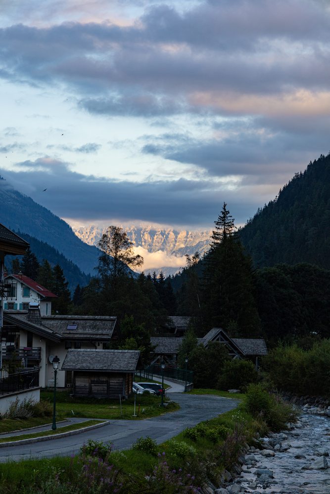 A stream and village houses leading towards mountains under evening clouds.