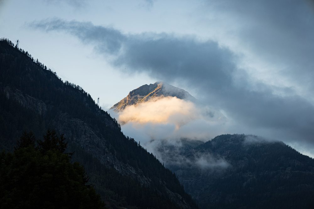 A mountain summit catching warm light through clouds above a dark valley.