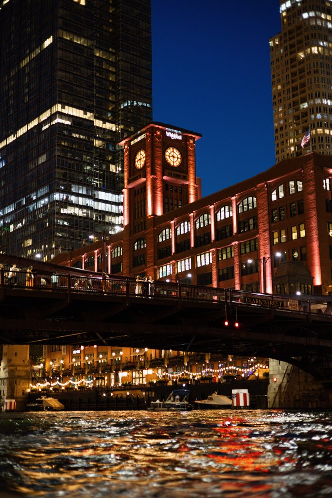 An illuminated brick clock tower above a bridge, with tall buildings behind and lights reflected in the river.