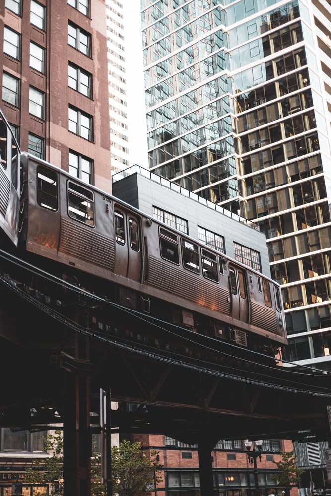 A silver train running above street level between tall city buildings.