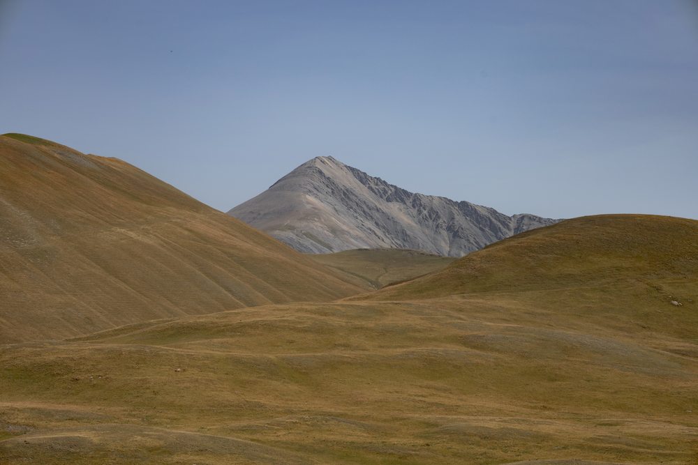 Rounded grassy hills lead to a grey triangular mountain peak beneath a blue sky.