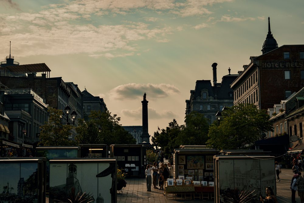 Art stalls and historic buildings surround a square, with a column silhouetted against a warm sky.