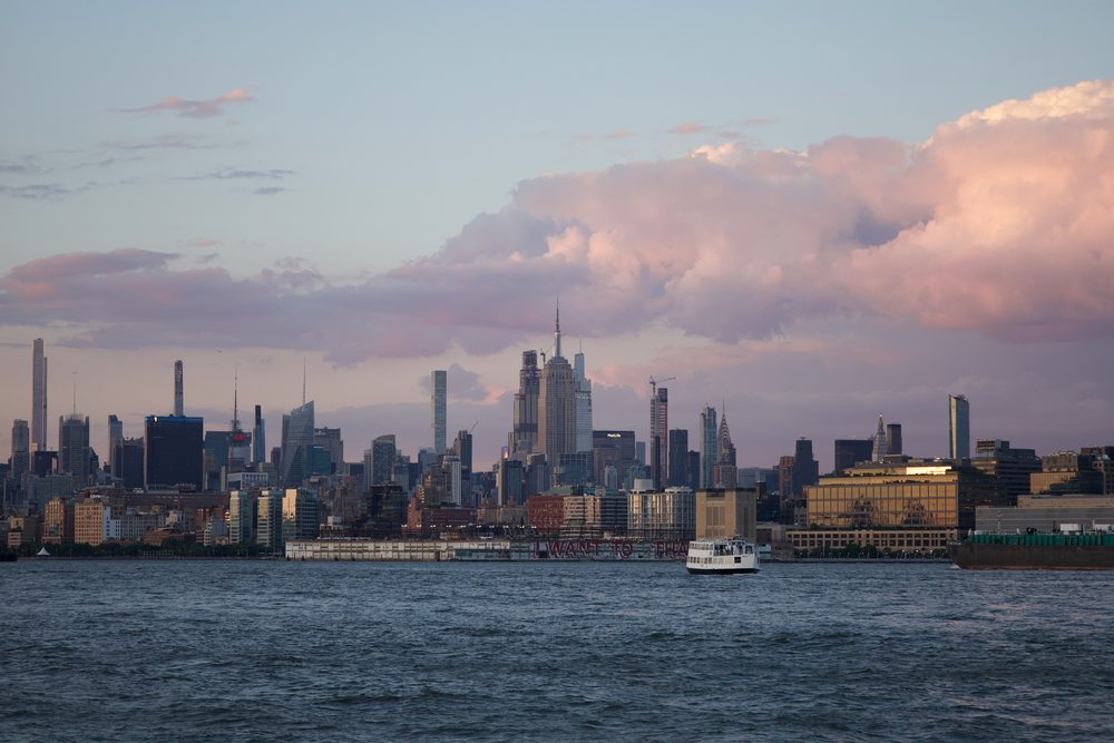 The Manhattan skyline below pink clouds, with a sailing boat on the water.