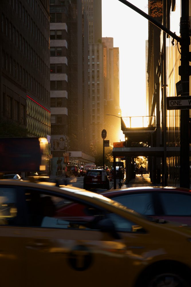 Sunlight falls between tall buildings as a yellow taxi crosses the foreground.