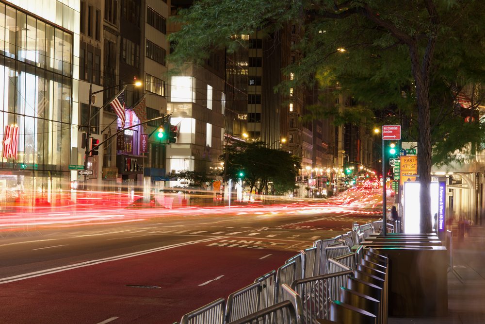 Long red light trails passing along a tree-lined city street at night.