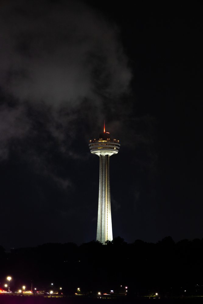 An illuminated observation tower rises into the dark sky, with a cloud drifting beside it.