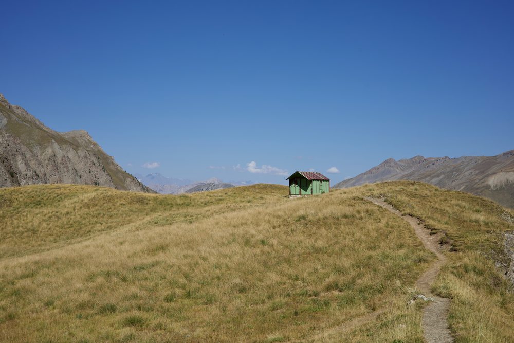 A small green shelter beside a path through an open alpine meadow.
