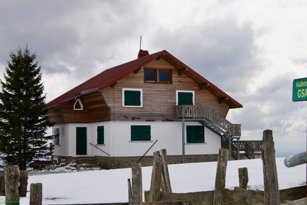A chalet with a red roof and green shutters stands in the snow behind a line of fence posts.