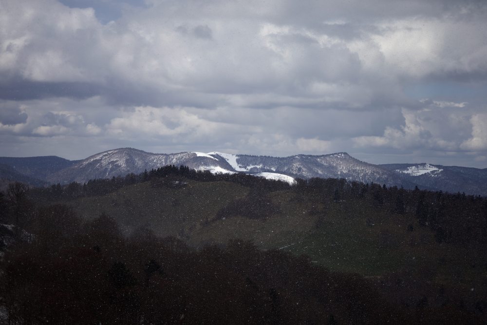 Snow falls across a white ridge above wooded hills under heavy clouds.