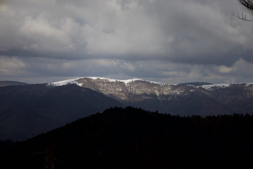 A sunlit snowy ridge above dark forested hills and beneath heavy clouds.